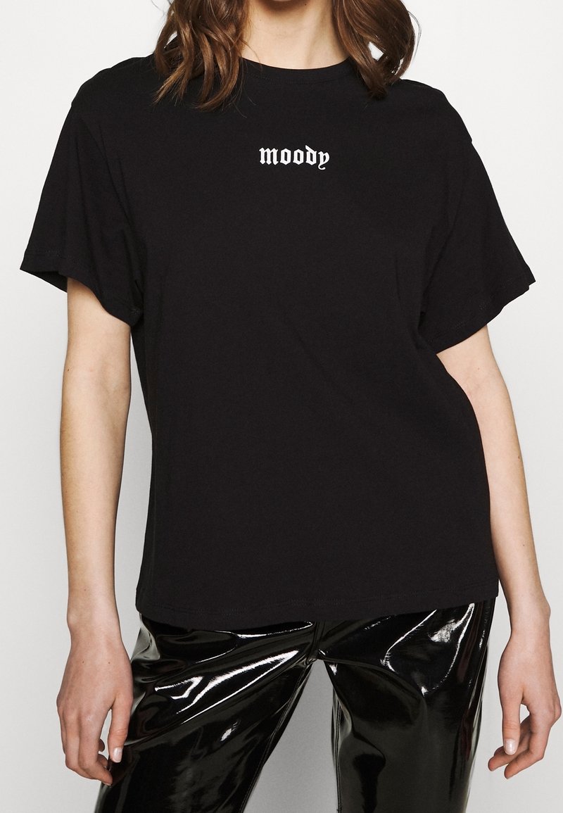 Femme portant un t-shirt noir avec le texte « moody » en police gothique blanche et un pantalon en vinyle noir brillant, sur un fond uni.
