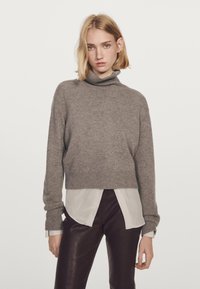Brązowy sweter z golfem wykonany z miękkiego dzianiny, z krojem skróconym i ściągaczami, nałożony na białą koszulę i brązowe spodnie.