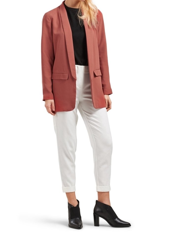 Femme portant un blazer couleur rouille, un haut noir, un pantalon blanc retroussé et des bottines noires à talons hauts, debout.