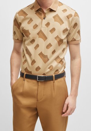 Homme portant un polo beige et marron à motifs, rentré dans un pantalon couleur chameau avec une ceinture noire, debout les mains détendues.