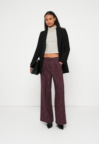 Zwarte blazer over een gestreepte grijze coltrui, gecombineerd met brede bordeauxrode broek met verticale witte strepen en kleine patronen. Zwarte handtas.