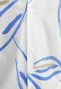 Camisa blanca con patrones abstractos de pinceladas en azul y beige, con un cierre de botones de plástico y una textura suave.