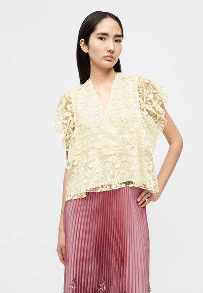 Κίτρινο τοπ από δαντέλα με φλοράλ μοτίβο και σχέδιο που δένει στο πλάι, με μανίκια τύπου φλάτερ. Συνδυασμένο με φούστα ροζ με πτυχώσεις, δημιουργώντας ένα στρωματοποιημένο λουκ.