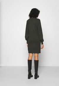 Abito maglione verde scuro con maniche lunghe a sbuffo; polsini e orlo a coste; indossato con stivali neri al ginocchio. Tessuto liscio, vestibilità rilassata.