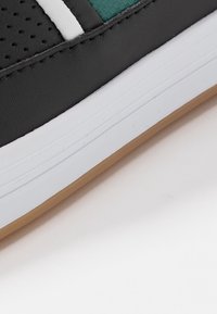 Zapatilla negra con diseño perforado, acentos en color teal, franjas blancas y suela de goma texturizada. Presenta una parte superior de sintético suave.