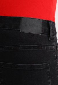 Černé džíny z denimu s hladkou texturou, se černou koženou etiketou na pase, oranžovým prošíváním a zadními kapsami bez viditelných dekorací.