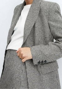 Černobílý kabát v vzoru psích zubů z texturované látky, s vykrojenými klopami, dvěma knoflíky a předními kapsami.