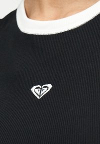 Κοντινό πλάνο μαύρου ριμπ πουκάμισου με λευκή στρογγυλή λαιμόκοψη και μικρό λευκό κεντημένο λογότυπο καρδιάς στο στήθος.