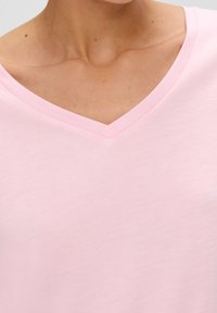 Detailní záběr na osobu oblečenou v světlém růžovém tričku s výstřihem do V, na kterém jsou vidět linie výstřihu a horní část hrudníku.