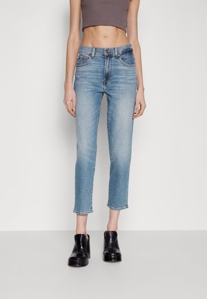 Vrouw draagt korte lichtblauwe spijkerbroek, zwarte plateau-laarzen en een mouwloze bruine crop top, staand tegen een effen witte achtergrond.