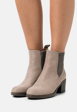 Bottines en suède beige avec des talons épais portées sur des jambes nues, présentant des panneaux latéraux élastiques noirs avec un motif en chevrons.