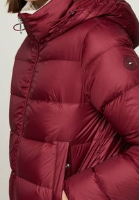 Bordowa pikowana kurtka puchowa z kapturem, z przeszyciami, srebrnymi elementami oraz naszywką z logo na rękawie.