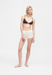 Γυναίκα όρθια, ξυπόλητη, φοράει μαύρο αθλητικό σουτιέν με σχέδιο στη ζώνη και λευκά σορτς ψηλόμεσα, μπροστά σε απλό λευκό φόντο.