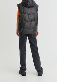 Veste matelassée noire avec capuche, présentant une texture lisse et brillante ainsi qu'un matelassage en chevrons. Associée à un pantalon gris foncé fuselé et des bottes épaisses noires.