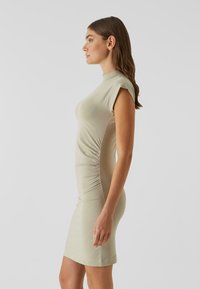 Tætsiddende kjole til knæene i blødt, lysebeige stof. Har høj hals og korte ærmer med rynkedetalje i siden for at skabe facon.