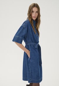 Denim-mekko keskisinisessä, lyhythihaisella, vyötäröllä vyöllä, sivutaskuilla, napituskiinnityksellä ja polvenpituiseen helmalla. Sileä pinta.
