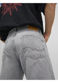 Vaqueros de mezclilla gris con corte clásico, que cuentan con dos bolsillos traseros y una parche de cuero texturizado en la cintura. Detalle de costuras sutiles visible.