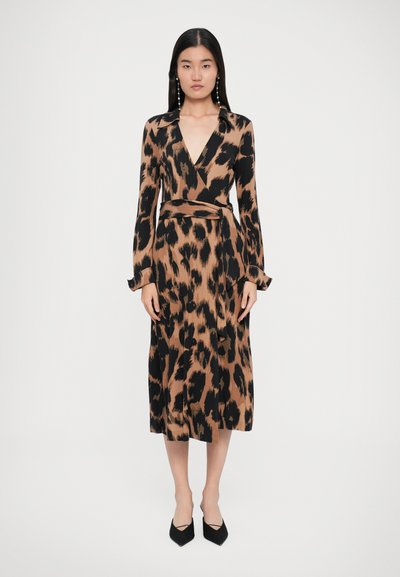 Femme aux longs cheveux noirs portant une robe portefeuille à imprimé animal marron et noir avec des manches longues et des chaussures noires à petit talon, se tenant devant un fond blanc.