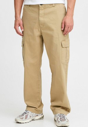 Beige cargo pants with a relaxed fit, featuring side pockets, a smooth texture, and reinforced stitching. Paired with gray sneakers.