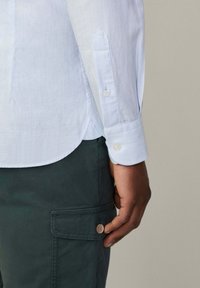 Lichtblauw gestreept shirt met lange mouwen, voorzien van knoopmanchetten. Draagt met donkergroene cargobroek met knoopzakdetail.