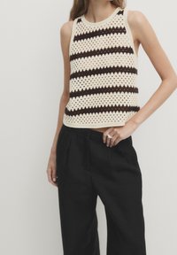 Donna che indossa un top in crochet senza maniche con righe orizzontali nere e crema e pantaloni neri larghi, con la mano appoggiata all'anca.