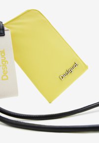 Geel synthetisch portemonnee met een glanzende afwerking, met een zilveren 'Desigual' logo, naast een beige stoffen riem. Zwarte textuurriem zichtbaar.