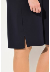 Marineblaues, tailliertes Kleid mit seitlichem Schlitz, aus einem strukturierten Stoff gefertigt. Der Saum ist gerade und das Kleid endet knapp über dem Knie.