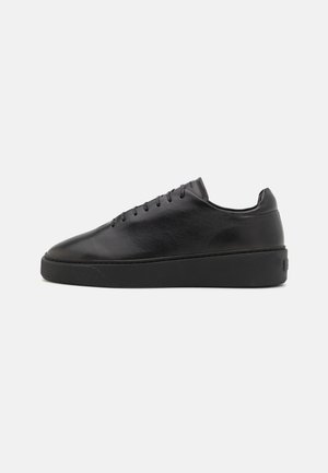 Czarny skórzany niskoprofilowy sneaker z pasującymi czarnymi sznurówkami i podeszwą, pokazany z profilu na jednolitym jasnym tle.