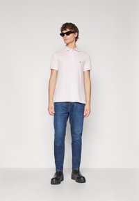 Polo de color rosa claro con cuello y tres botones, combinado con jeans de mezclilla azul oscuro y zapatos negros gruesos. Los accesorios incluyen gafas de sol.