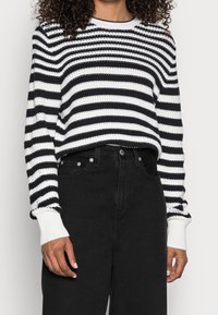 Sweter dzianinowy w czarno-białe paski z długimi rękawami, z prążkowanymi mankietami i okrągłym dekoltem; zestawiony z czarnymi dżinsami z wysokim stanem.
