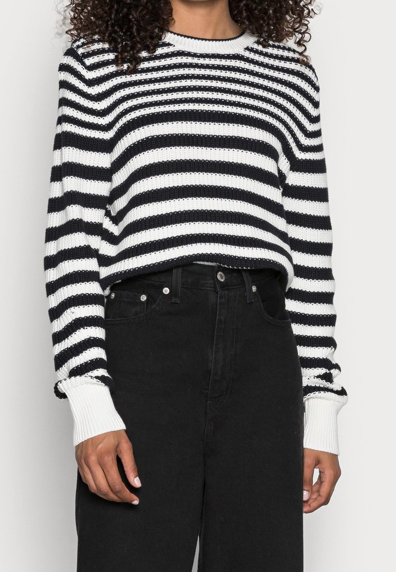 Sweter dzianinowy w czarno-białe paski z długimi rękawami, z prążkowanymi mankietami i okrągłym dekoltem; zestawiony z czarnymi dżinsami z wysokim stanem.