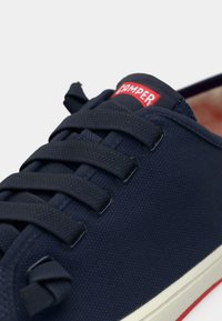 Σκουρόχρωμο ναυτικό πάνινο παπούτσι με υφή στην επιφάνεια, στρογγυλεμένη μύτη, λευκή σόλα σε αντίθεση και κόκκινο λογότυπο "Camper" στη γλώσσα.