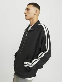Sort zip-up jakke med hvide striber på ærmerne, der har en afslappet pasform, to forlommer og et glat tekstureret stof.
