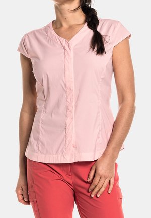 Woman wearing a light pink short-sleeve blouse and coral pants, standing with one hand resting on thigh against a plain background.