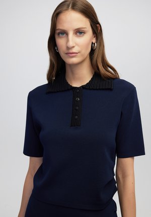 Mujer de piel clara y cabello castaño claro, vestida con una blusa de manga corta azul marino que tiene un cuello de canalé negro y una fila de botones, de pie frente a un fondo liso.