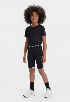 Kind mit lockigem Haar, trägt schwarze Nike Pro Shorts, schwarzes T-Shirt, schwarze Socken und weiße Nike-Sneaker, steht vor einem schlichten Hintergrund.