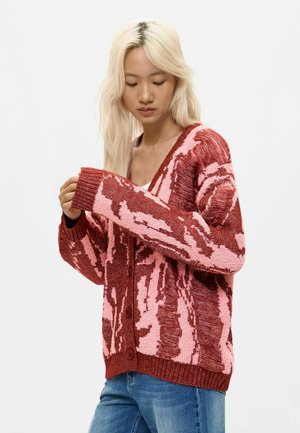Frau mit langen blonden Haaren, die eine rot-rosa gemusterte Strickjacke und blaue Jeans trägt, sieht nach unten und richtet ihren Ärmel vor einem einfarbigen Hintergrund.