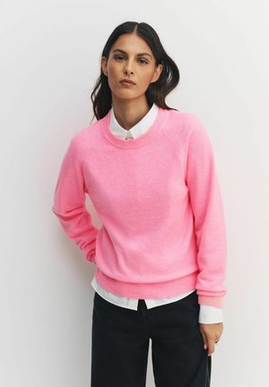 Pink sweatshirt with ribbed cuffs and hem, featuring a round neckline and raglan sleeves, layered over a white shirt, paired with dark pants.