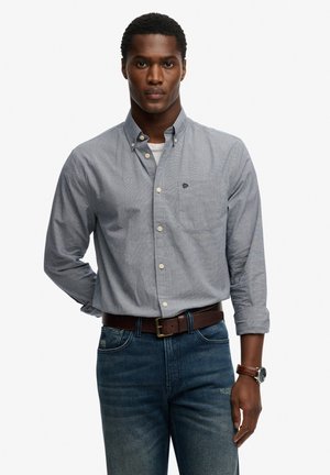 Camisa de cuadros gris y blanca con botones, mangas largas, con bolsillo en el pecho y logo bordado, combinada con vaqueros azules y un cinturón marrón.