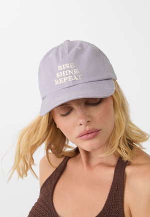 Helle lavendelfarbene Baumwollmütze mit gesticktem Text "RISE SHINE REPEAT" in Creme. Sie hat einen gebogenen Schirm und einen verstellbaren Rückenriemen.