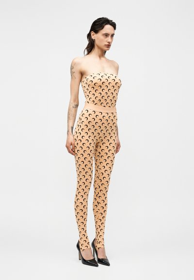 Ženska v bež kombinezonu brez naramnic s črnimi polmeseci, stoji v črnih ostrih visokih petah pred enobarvnim ozadjem.