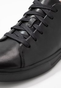 Gros plan d'une basket en cuir noir avec des lacets noirs passés à travers cinq œillets de chaque côté, reposant sur une surface blanche.