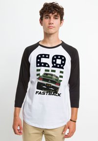 Baseball T-shirt med hvid krop og sorte raglanærmer. Har grafik af en bil, teksten "68 Highland Green FASTBACK," grønne striber og stjerner.
