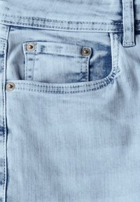 Tissu en denim bleu clair présentant une poche avant avec des rivets en métal couleur cuivre et des motifs de décoloration subtils pour la texture.