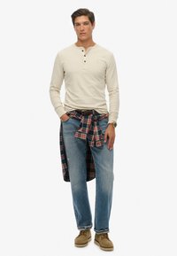 Henley beige de manga larga con tres botones, combinado con jeans de mezclilla azul claro. Camisa a cuadros roja y azul marino atada a la cintura, calzado marrón.