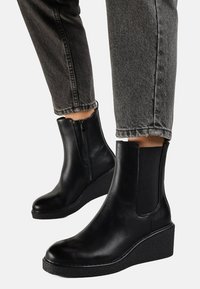 Bottines noires en cuir lisse avec un semelle en wedge. Elles sont dotées de panneaux élastiques sur les côtés et d'une fermeture éclair sur le côté, posées sur un jean gris.