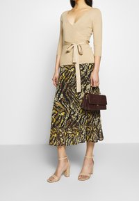 Beige ribbat inlindad topp med knytband i midjan, ihopparad med en mönstrad brun och gul midikjol, samt en texturerad burgundy-handväska. Beige klacksandaler.