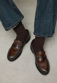 Bruine leren loafers met een textuur van bruin bontpaneel, zwarte zool en dubbele banddetail, gecombineerd met donkerbruine sokken en blauwe spijkerbroek.