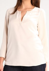 Donna che indossa una blusa a maniche lunghe di colore crema con scollatura a V e pantaloni scuri, tagliati alle spalle e ai fianchi.