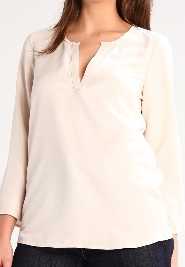 Donna che indossa una blusa a maniche lunghe di colore crema con scollatura a V e pantaloni scuri, tagliati alle spalle e ai fianchi.